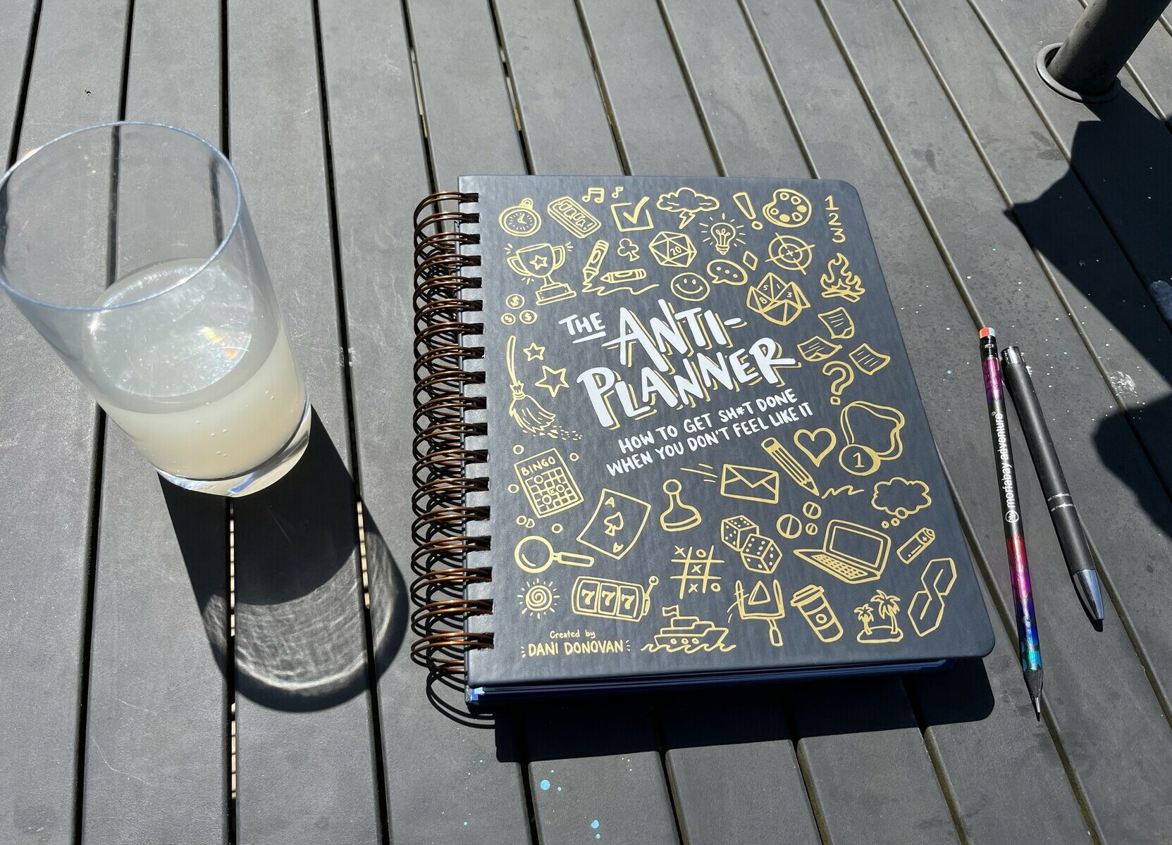 A large, ring bound planner with a pencil and a glass of ginger beer lay on my garden table in the sun