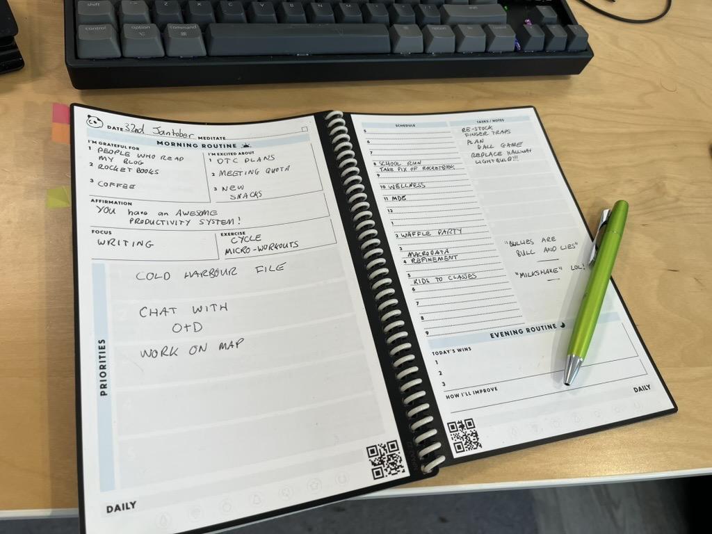 My RocketBook Panda Planner is lying open on my desk with my keyboard above it and a green pen resting on it.

It says I’m excited about “OTC Plans, meeting quota, and new snacks”.

It says my priorities are the “Cold Harbour” file, “Chat with O and D”, and “Work on Map”.

My schedule for the day includes Wellness, MDE, Waffle Party, and Macrodata Refinement.

My tasks and notes include “Re-stock finger traps”, “Plan Ball Game”,“Replace Hallway Lightbulb” and “Milkshake, lol”.

There is a quote written down: “Bullies are Bull and Lies”