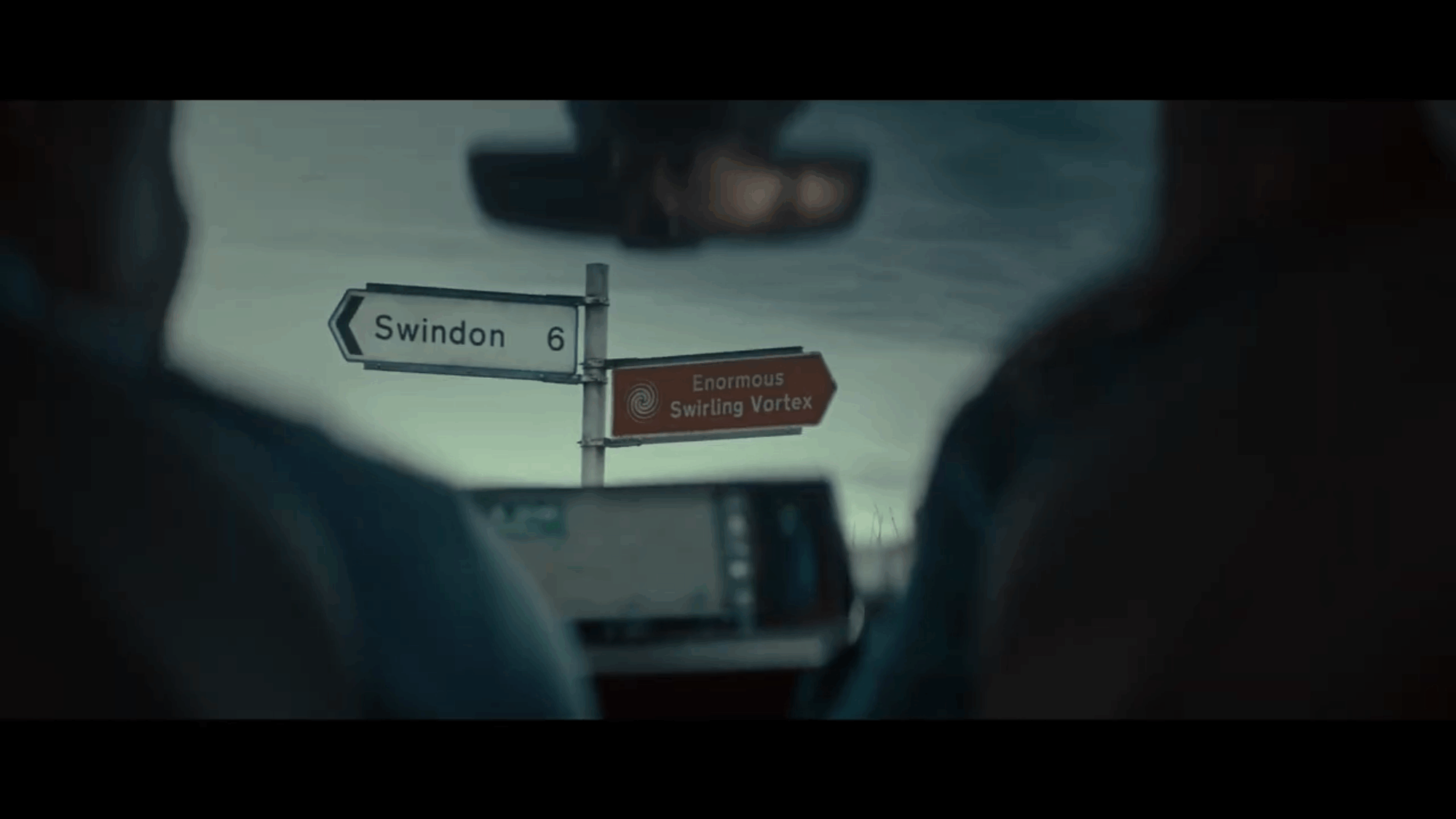 A screenshot of the ad mentioned. The camera is inside a car looking forward out of the windscreen. There is a direction sign in front of the car. The left arrow says it goes to Swindon. The right arrow says it goes to “Enormous Swirling Vortex”
