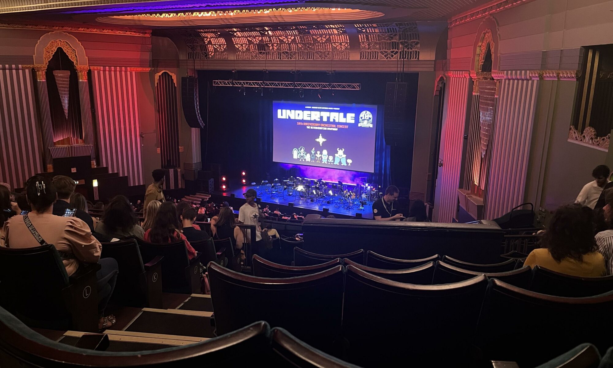 A view from high up in a theatre. A screen shows some video game characters and the text “Undertale: 10th Anniversary Orchestral Concert. The determination symphony”