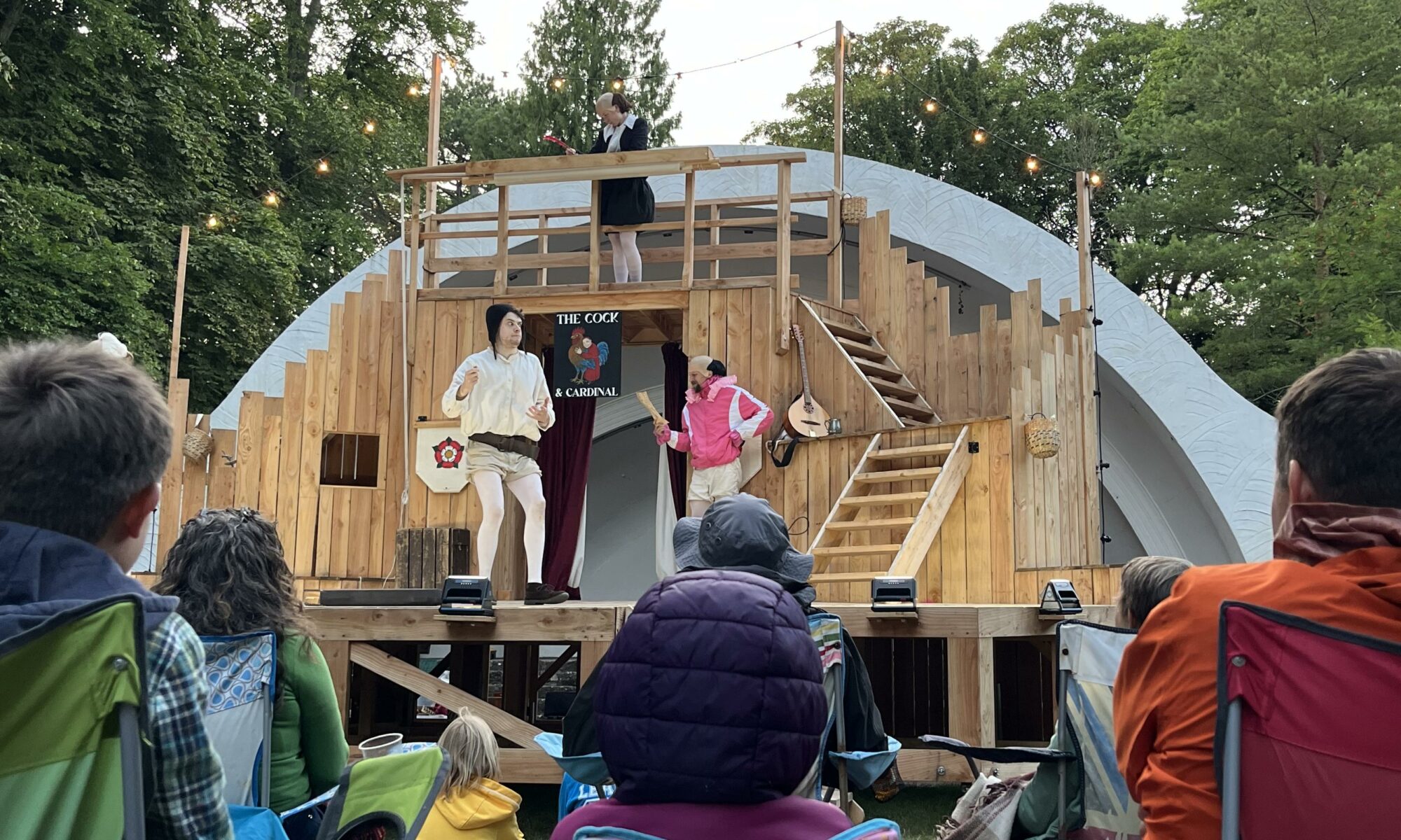 A wooden outdoor stage surrounded by trees. On the stage are theee actors. One in bright pink with white bloomers and tights. One in all white. And one in white tights, black bloomers, a black jacket and wearing a comedy bald headpiece and writing with a quill. They are all in amusing poses. People in camping chairs are in the foreground.