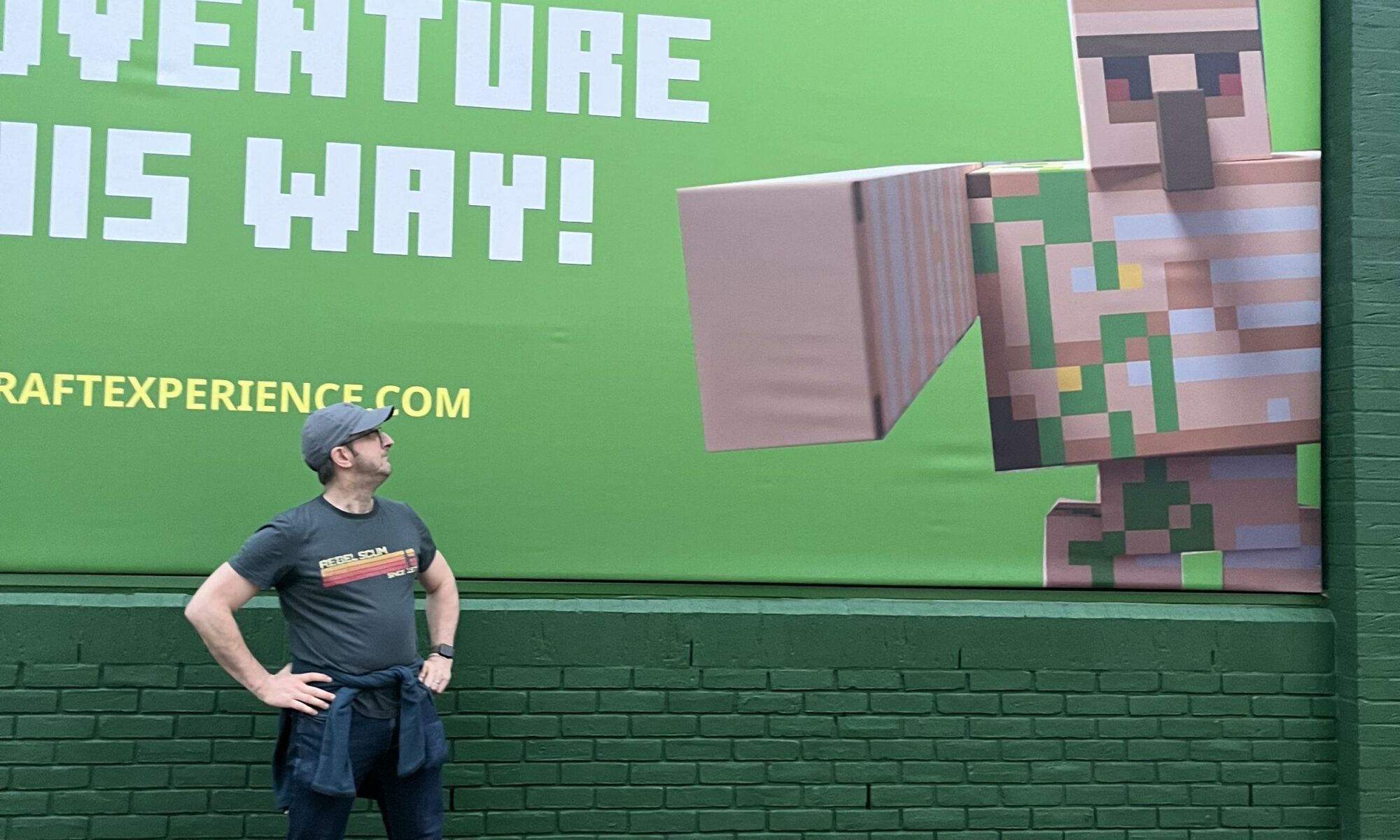 Ross stands in front of a large poster reading “Adventure this way!” that has an Iron Golem on it that is pointing to where Ross is standing.