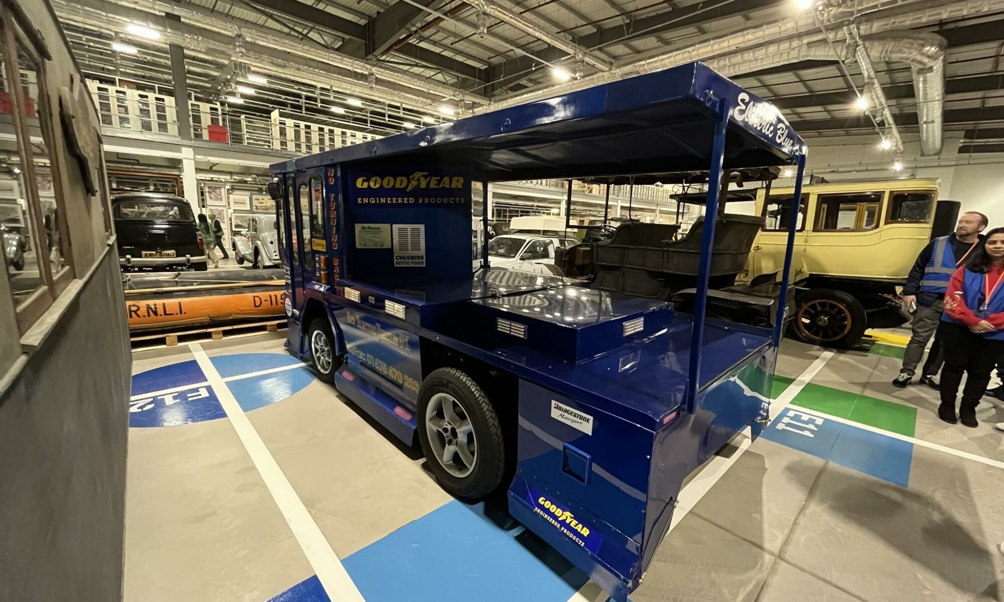 A shiny blue milk float sits among other old vehicles in a large warehouse.