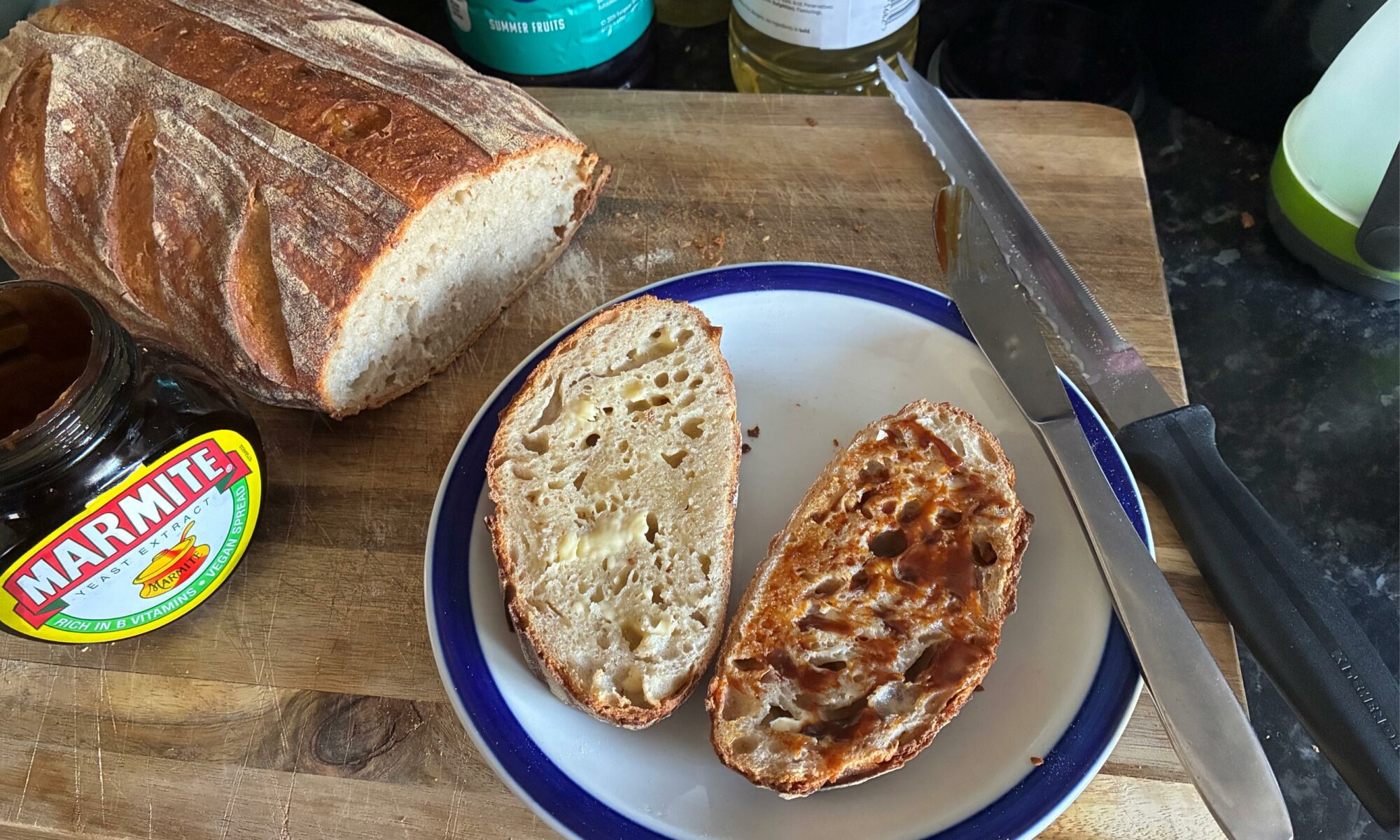 A bread board with a freshly-baked sourdough loaf, a plate with two slices of that bread, one has butter, one has butter and marmite. The marmite jar is also in view.